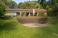 Außenansicht des Pfarrkindergartens Neubeuern, ein modernes Gebäude mit heller Fassade und großen Fenstern. Im Vordergrund sind ein grüner Rasen und einige Spielgeräte zu sehen. Das Gebäude ist von Bäumen umgeben, und der Himmel ist klar.