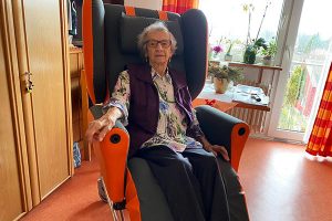 Eine ältere Dame in einem orange-schwarzen Pflegestuhl in ihrem Zimmer.