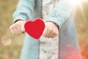 Ein kleines Kind, dessen Kopf nicht zu sehen ist, hält ein rotes Holzherz.