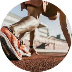 Ein Sprinter in Sportkleidung und orangenen Laufschuhen steht in Startposition auf einer Laufbahn. Die Aufnahme ist aus Bodennähe gemacht, sodass hauptsächlich die Beine und Füße im Fokus stehen, während im Hintergrund unscharf das Stadion zu erkennen ist.