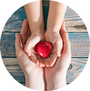 Im Hintergrund sind blaue Holzlatten zu sehen. Darauf liegen die Hände einer Erwachsenen Person, die die beiden Kinderhände halten. Dieses wiederrum hält ein rotes Herz in der Hand.