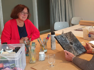 Eine Frau mit roter Strickjacke sitzt an einem Tisch auf dem Pinsel, Farben und zwei Arme, die ein Bild halten, zu sehen sind.