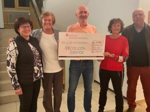 Fünf Erwachsene stehen nebeneinander und lächeln in die Kamera. Die mittlere Person hält einen großen symbolischen Scheck der Sparkasse Rosenheim-Bad Aibling. Auf dem Scheck steht: „PROTEGONA SPENDE, 1.000 €, am 24.11.2023“. Im Hintergrund sind eine Treppe und ein Geländer zu sehen.